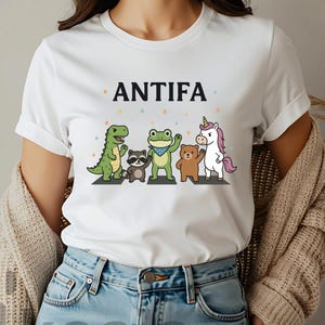 May include: White t-shirt with the word "ANTIFA" above a graphic of cartoon animals crossing a street. The animals include a green dinosaur, raccoon, frog, bear, and unicorn. The shirt is being worn by a person wearing blue jeans and a beige cardigan.