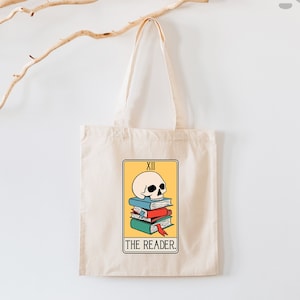 May include: A white tote bag with a yellow and white illustration of a skull on top of a stack of books. The illustration is labeled "XII THE READER."