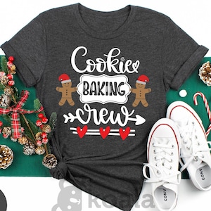 May include: Dark gray heather t-shirt with the text "Cookie Baking Crew" and gingerbread man graphics. The shirt is styled with white sneakers, candy canes, and Christmas decorations.