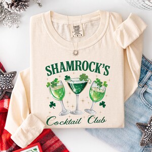 May include: Ivory long-sleeve shirt with the text "SHAMROCK'S Cocktail Club" in green. The design features three green cocktails with shamrocks. The shirt is laid flat with a pair of blue jeans and a star-shaped decoration.