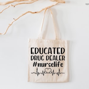 May include: A white canvas tote bag with black text that reads "Educated Drug Dealer #nurselife" and a black and white heartbeat graphic with a heart in the middle.