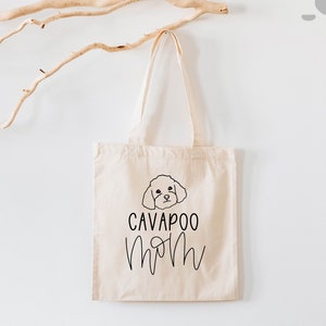 May include: A beige canvas tote bag with a black Cavapoo dog illustration and the words "CAVAPOO MOM" in a handwritten font. The bag hangs against a white wall with a light brown branch.