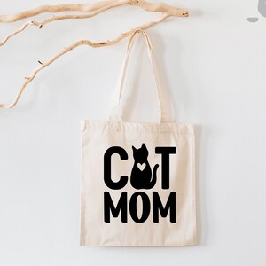 Peut inclure: Un sac fourre-tout en toile blanche avec du texte noir qui dit "CAT MOM" et une silhouette noire d'un chat avec un c&oelig;ur sur la poitrine.