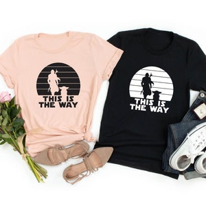 May include: Two t-shirts with a black and white silhouette of a character from the Star Wars franchise, with the text "This is the way" printed on each shirt. One shirt is heather peach and the other is black.