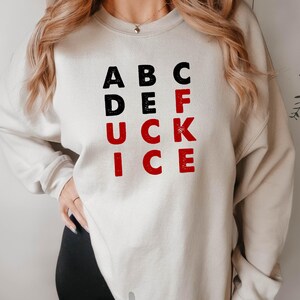 May include: A sand-colored sweatshirt with the text "ABC DE F UCK ICE" in black and red block letters. The sweatshirt is a crew neck style and has long sleeves. The image also shows black shorts.