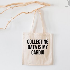 K&ouml;nnte beinhalten: Eine wei&szlig;e Einkaufstasche mit schwarzem Text, der "COLLECTING DATA IS MY CARDIO" lautet.