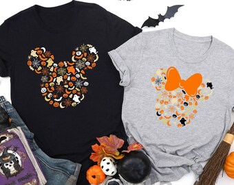 May include: Black and light gray t-shirts with a Halloween-themed design. The design on each shirt is a Mickey Mouse head shape filled with Halloween icons like ghosts, pumpkins, and spiderwebs. The light gray shirt has an orange bow.