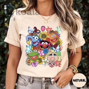 May include: A light beige t-shirt with a colorful graphic print of Sesame Street characters, including Big Bird, Cookie Monster, Elmo, and Miss Piggy, surrounded by flowers.