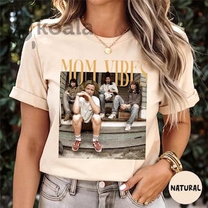 May include: A beige t-shirt with a graphic print of four people sitting on a porch. The graphic includes the text "MOM VIBES".