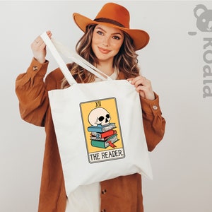 May include: White tote bag with a yellow and orange tarot card design featuring a skull on top of a stack of books. The card reads "XII THE READER".