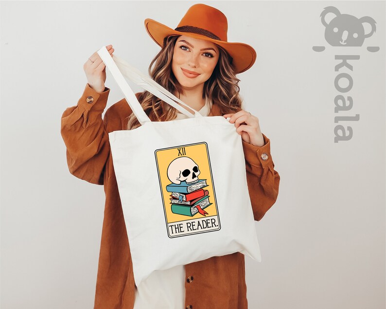 May include: White tote bag with a yellow and orange tarot card design featuring a skull on top of a stack of books. The card reads "XII THE READER".