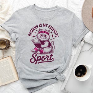 a gray t-shirt with a graphic design, a hat, a cup of coffee, a book, and a clock, all arranged on a white surface.