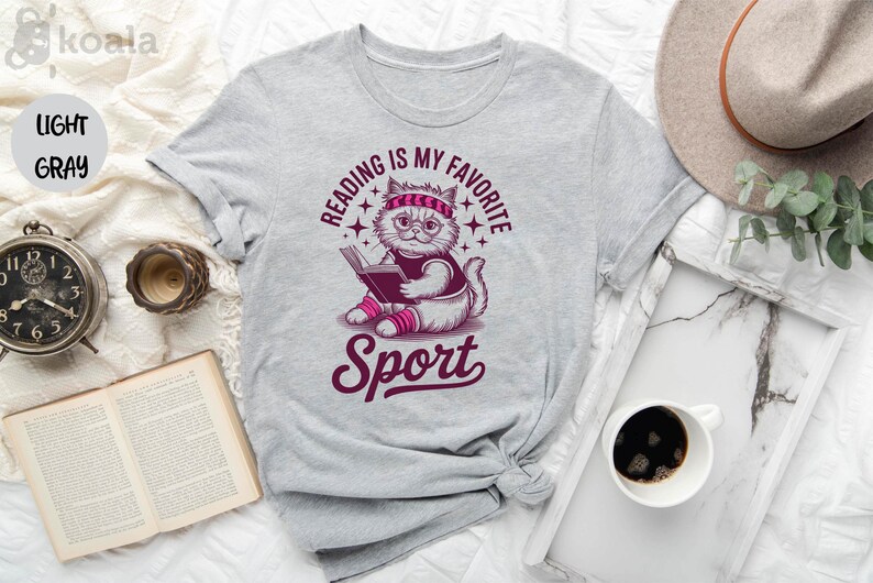 a gray t-shirt with a graphic design, a hat, a cup of coffee, a book, and a clock, all arranged on a white surface.