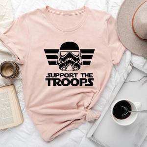 Peut inclure: Un t-shirt rose avec un graphique noir d'un casque de Stormtrooper de Star Wars avec le texte "SUPPORT THE TROOPS" en dessous.