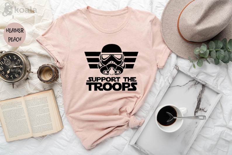 Peut inclure: Un t-shirt rose avec un graphique noir d'un casque de Stormtrooper de Star Wars avec le texte "SUPPORT THE TROOPS" en dessous.