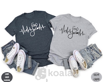 May include: Two t-shirts with a heartbeat graphic and a stethoscope with the letters CNA. The shirt on the left is dark heather gray and the shirt on the right is light gray.