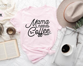 May include: A pink t-shirt with the text "Mama needs coffee" in black cursive lettering. The shirt is knotted and laid out on a white surface with a book, a clock, a cup of coffee, a hat, and a plant.