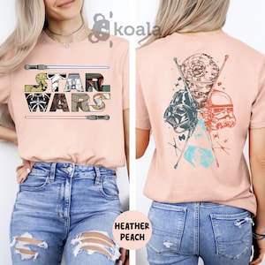 May include: A heather peach t-shirt with the text "STAR WARS" on the front and a graphic of Star Wars characters on the back. The shirt is being modeled by a person wearing blue jeans.