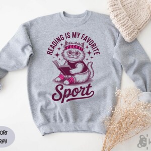 A gray sweatshirt with a graphic design of a cat reading a book, accompanied by the text "Reading is my favorite Sport" and "Sport" written below the cat.