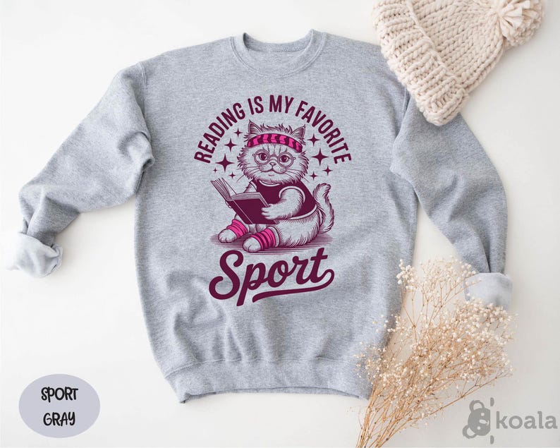 A gray sweatshirt with a graphic design of a cat reading a book, accompanied by the text "Reading is my favorite Sport" and "Sport" written below the cat.