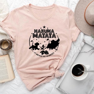 May include: A pink t-shirt featuring a black graphic print of characters from the film The Lion King. The graphic includes the text "Hakuna Matata" and "It means no worries".