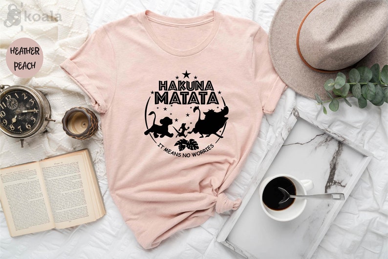 May include: A pink t-shirt featuring a black graphic print of characters from the film The Lion King. The graphic includes the text "Hakuna Matata" and "It means no worries".