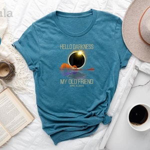 May include: A teal blue t-shirt with the text "Hello Darkness My Old Friend" and the date "April 8, 2024". The design features a stylized image of a solar eclipse.