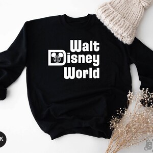May include: Black sweatshirt with the words "Walt Disney World" in white block letters. The design includes a Mickey Mouse head and a globe. A white knit beanie and dried flowers are in the background. The word "BLACK" is in a black circle.