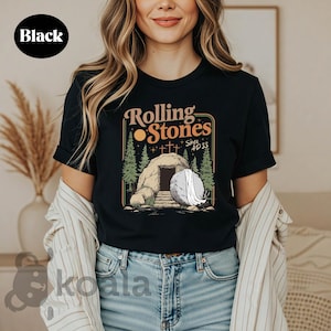 May include: Black t-shirt featuring a graphic with the text "Rolling Stones Since AD 33". The design includes a tomb, trees, and a rising sun. The shirt is a classic crew neck.