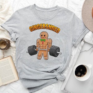 May include: Light gray t-shirt featuring a cartoon gingerbread man lifting weights. The text "Gingershred" is arched above the gingerbread man. The gingerbread man has a green bow tie and muscular arms. The barbell has black weights.