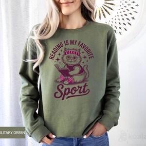 A woman wearing a green sweatshirt with a graphic design of a cat reading a book and the text "Reading is my favorite sport" printed on it.