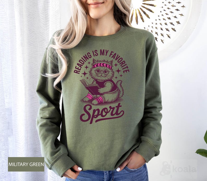 A woman wearing a green sweatshirt with a graphic design of a cat reading a book and the text "Reading is my favorite sport" printed on it.
