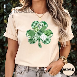 May include: Beige t-shirt featuring a large, patchwork shamrock design in various shades of green. The shamrock is composed of different patterns, including gingham, floral, and paisley. The shirt has short sleeves and a crew neck.