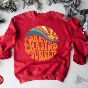 May include: Red sweatshirt with a retro-style graphic of a sun setting over a wave. The graphic says "FOREVER CHASING SUNSETS".