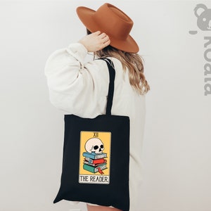May include: Black tote bag with a yellow and black tarot card design. The card depicts a skull on top of a stack of books with the text "THE READER" and "XII" on the card.