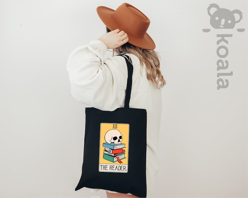 May include: Black tote bag with a yellow and black tarot card design. The card depicts a skull on top of a stack of books with the text "THE READER" and "XII" on the card.