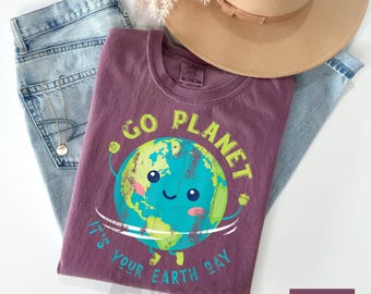 May include: A maroon t-shirt with a graphic of a smiling Earth holding a sign that says "Go Planet It's Your Earth Day".