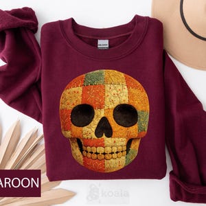 May include: Maroon sweatshirt featuring a colorful patchwork skull design. The skull has black eye sockets and a detailed patchwork pattern in shades of orange, yellow, green, and red. The word "MAROON" is displayed in a white font on a maroon background.