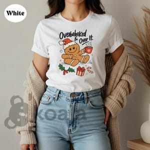 May include: White t-shirt featuring a gingerbread man wearing a Santa hat and holding a mug. The shirt has the text "Overbaked & Over It." Festive design includes holly, a gift, and a candy cane. A cozy holiday themed tee.