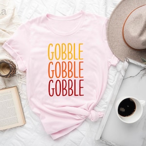 Op de afbeelding: Een lichtroze t-shirt met het woord "GOBBLE" in grote, kleurrijke letters. Het woord is drie keer gestapeld, met kleuren die overgaan van geel naar oranje naar rood. Een neutrale hoed en een dienblad met koffie zijn ook zichtbaar.