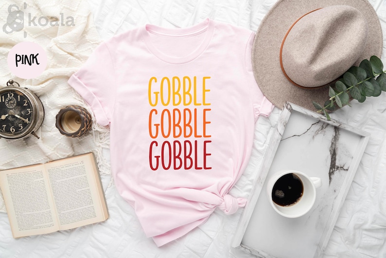 Op de afbeelding: Een lichtroze t-shirt met het woord "GOBBLE" in grote, kleurrijke letters. Het woord is drie keer gestapeld, met kleuren die overgaan van geel naar oranje naar rood. Een neutrale hoed en een dienblad met koffie zijn ook zichtbaar.