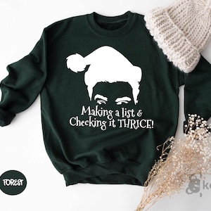May include: Forest green sweatshirt with a white graphic of a person wearing a Santa hat and the text "Making a list & Checking it THRICE!". A cream-colored knit hat and dried flowers are next to the sweatshirt.