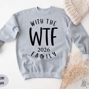 A gray sweatshirt with the text "WITH THE WTF 2026 FAMILY" printed on it, along with a knit hat and some dried flowers.