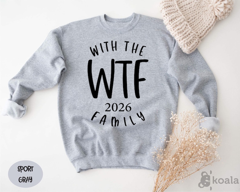 A gray sweatshirt with the text "WITH THE WTF 2026 FAMILY" printed on it, along with a knit hat and some dried flowers.