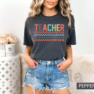 May include: A dark gray t-shirt with the word "TEACHER" in rainbow colors and a checkered pattern. The text "BELIEVER", "EDUCATOR", "INNOVATOR", and "MOTIVATOR" are listed below the word "TEACHER" in a smaller font.