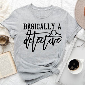 May include: A light gray t-shirt with the text "BASICALLY A detective" in black lettering. A magnifying glass is included in the text design.