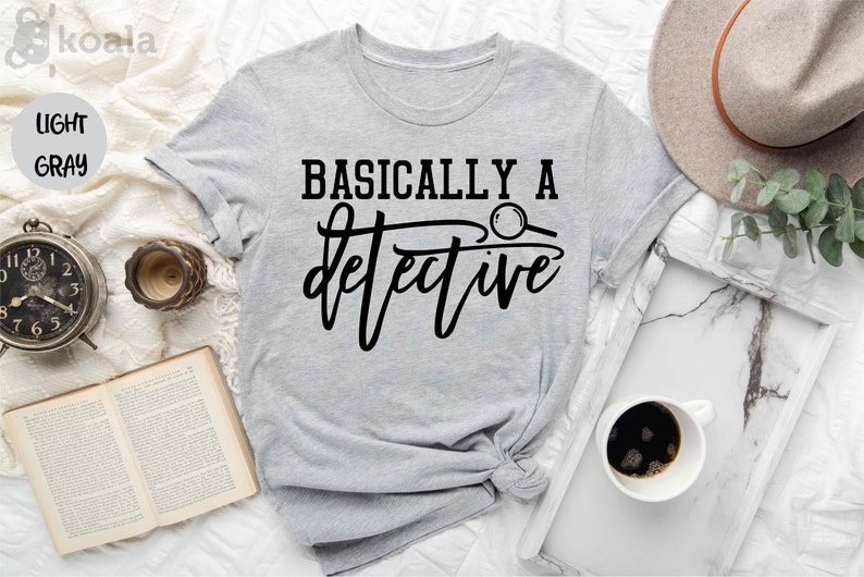 May include: A light gray t-shirt with the text "BASICALLY A detective" in black lettering. A magnifying glass is included in the text design.