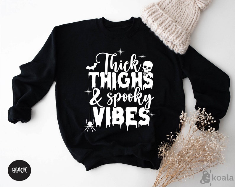 Op de afbeelding: Zwart sweatshirt met de witte tekst "Thick Thighs & Spooky Vibes". Het ontwerp bevat een vleermuis, schedel en spin. Een cr&egrave;mekleurige gebreide muts is zichtbaar. Het woord "BLACK" staat in een zwarte cirkel linksonder.