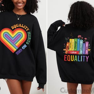 May include: Black sweatshirt with a rainbow heart and the text "Equality Hurts No One". The back of the sweatshirt has a stack of books with words like "Love", "Unity", and "Change" and the text "Equality" in rainbow colors.