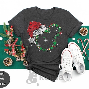 May include: Dark gray heather t-shirt featuring a Christmas-themed Mickey Mouse design. The design includes a Santa hat, a Mickey Mouse head shape, and festive colors. White sneakers and holiday decorations complete the scene.